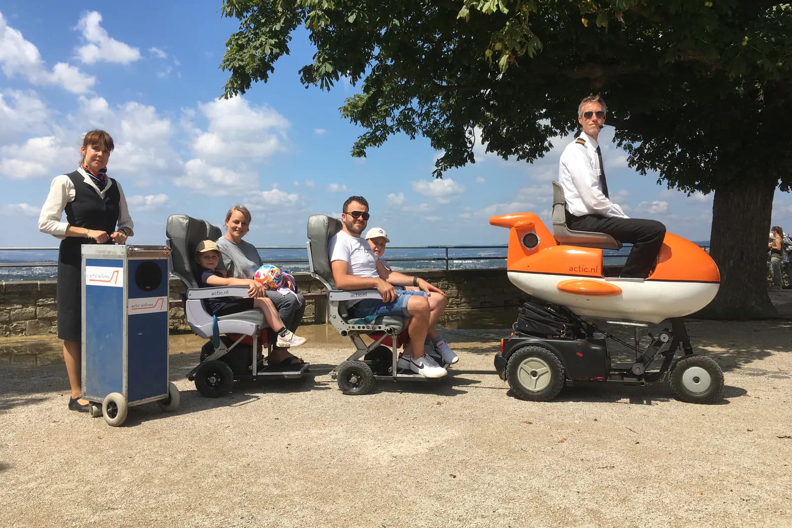 Een stewardess duwt een bagagetrolley naast twee rijen vliegtuigstoelen op wielen met passagiers erin, met vooraan een piloot op een oranje mini-vliegtuigje.