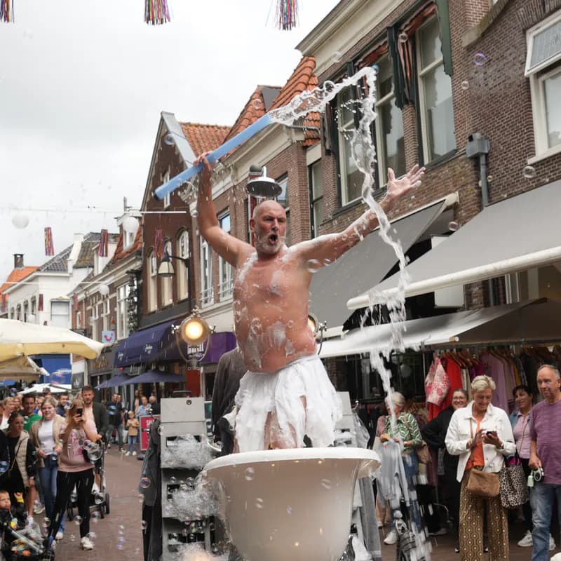 Dezelfde badgast staat rechtop in het bad in een winkelstraat, een blauwe staaf omhoog in zijn hand, terwijl het water in een boog naast hem omlaag valt en het winkelend publiek blijft kijken.