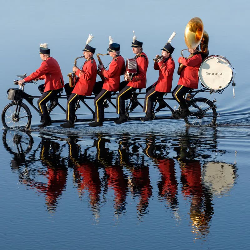 De tandem rijdt over spiegelglad water, met de zes muzikanten en hun instrumenten scherp weerspiegeld in het wateroppervlak en de tekst fietsorkest.nl op de grote trom.