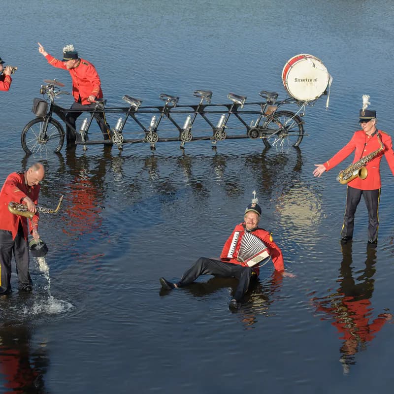 In ondiep water rond de stilstaande tandem giet een muzikant water uit zijn schoen, terwijl een ander languit in het water zit en accordeon speelt.