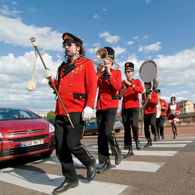 Het korps steekt achter elkaar een zebrapad over, een tamboer-maitre met witte handschoenen en staf voorop, terwijl een rode auto voor het zebrapad wacht.